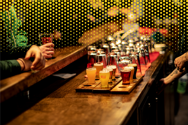 A wooden bar showing a several flights of beer being served. In the background, clean, empty Candid Brewing glasses. A man rests his elbows on the bar as a server's arms can be seen reaching under the bar. 