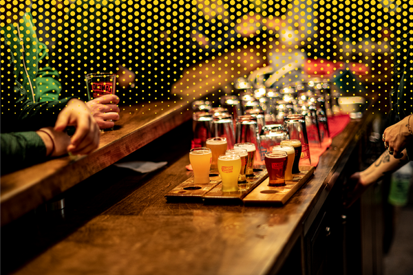 A wooden bar showing a several flights of beer being served. In the background, clean, empty Candid Brewing glasses. A man rests his elbows on the bar as a server's arms can be seen reaching under the bar.