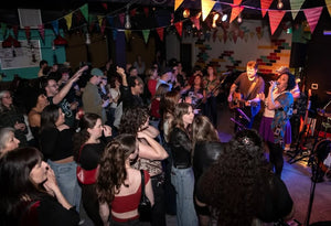 A crowded indoor band performance space with people dancing and singing along to music played from a stage decorated with colorful triangular flags.