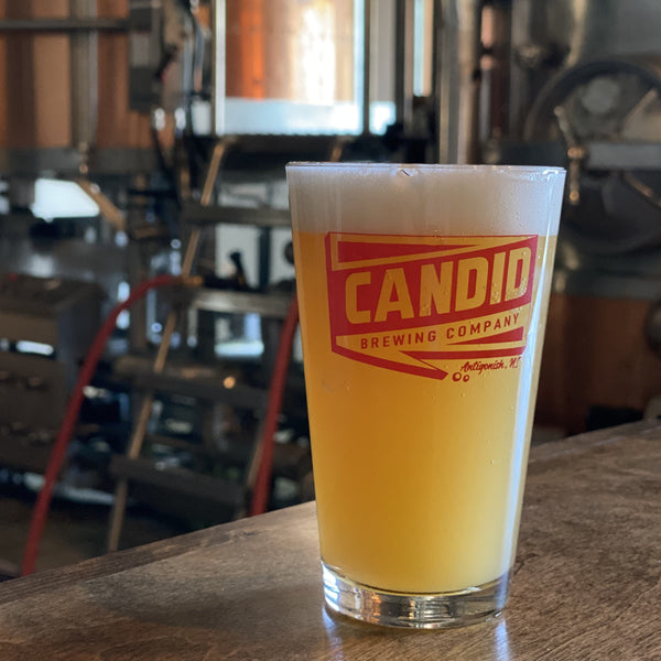 A Candid branded pint glass with a hazy beer in it. In the background, brewing equipment is shown. 