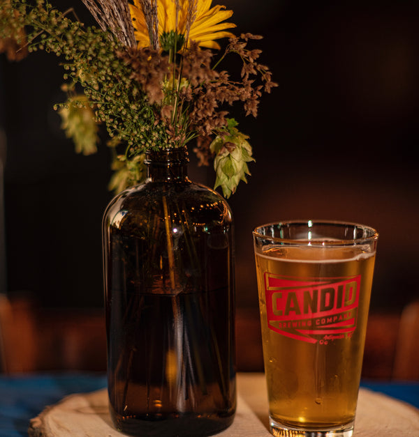 An image of a Candid branded pint glass filled with beer and next to a vase filled with flowers.